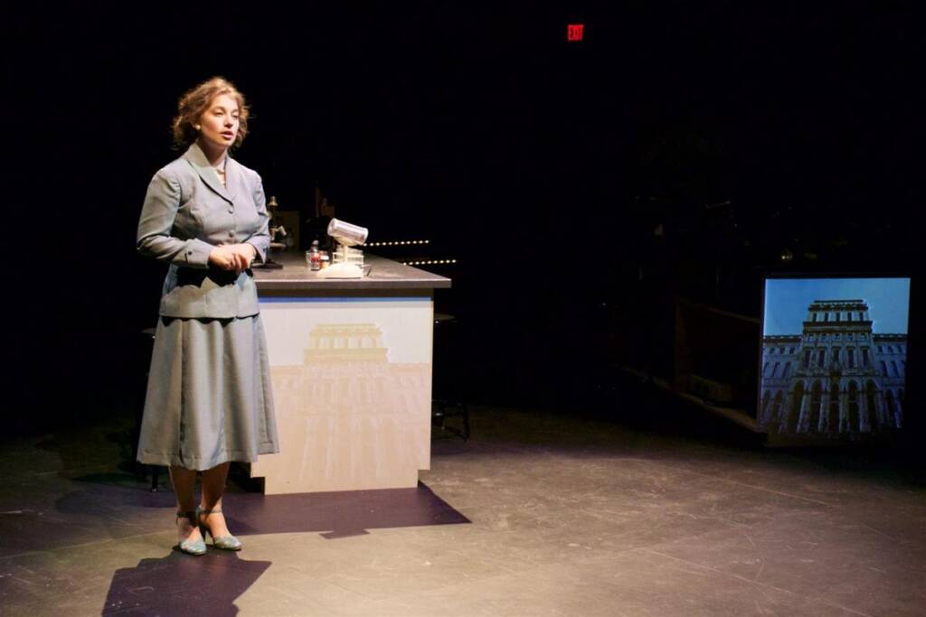 A woman in a vintage outfit stands on a dimly lit stage beside a desk with a microphone. A projected image of a building is visible on a screen to the right. The background is dark, highlighting her presence under the spotlight.