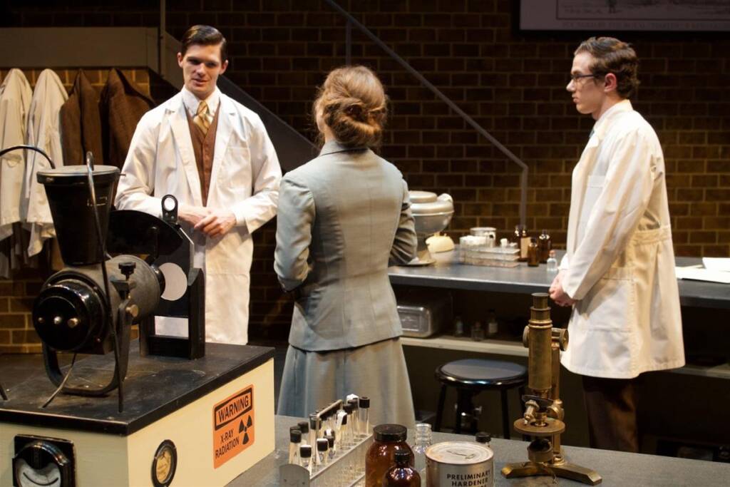 Three people in a vintage laboratory setting, all wearing lab coats. They are surrounded by scientific equipment, including a microscope and a Geiger counter with a warning label. A woman in a green outfit faces two men. The background features a brick wall.