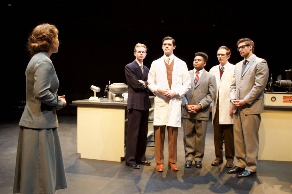 A group of people in formal attire is standing on a stage resembling a laboratory setting. One person, wearing a lab coat, faces another in a suit across a counter with scientific equipment. The others observe attentively.