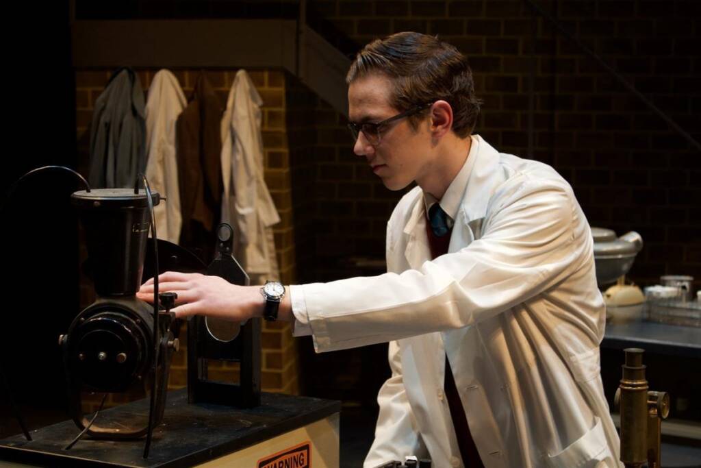 A person in a white lab coat and glasses operates a machine in a laboratory setting. Lab coats hang in the background. The person appears focused, adjusting dials on the equipment. The setting suggests a scientific or research environment.