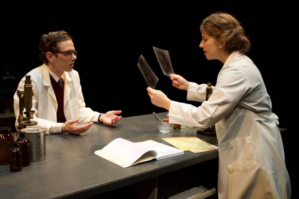 Two scientists in lab coats are engaged in a discussion at a lab table. One is seated with outstretched hands, and the other is standing, holding up two X-ray films. A microscope, bottles, and open book are on the table.