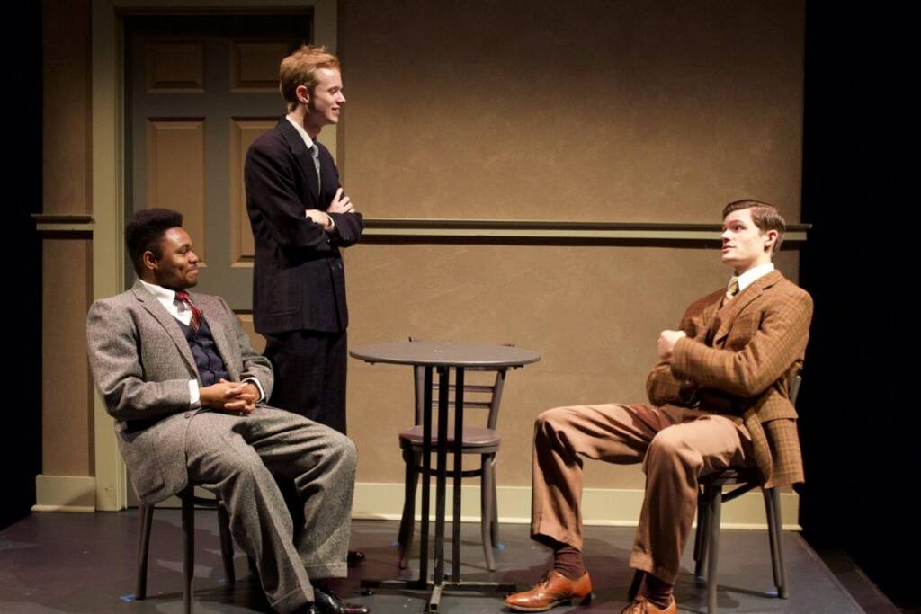 Three men wearing suits are in a room with neutral-colored walls. Two are seated at a small round table, while one stands nearby, smiling with his arms crossed. The setting resembles a stage play or theatrical production.