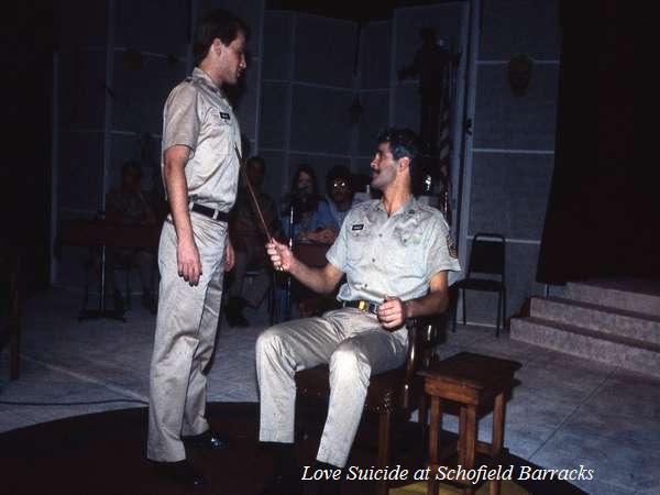 Two men in military uniforms are on a dimly lit stage. One is standing, while the other is seated holding a stick. The scene appears to be part of a theatrical performance. The text "Love Suicide at Schofield Barracks" is visible at the bottom.