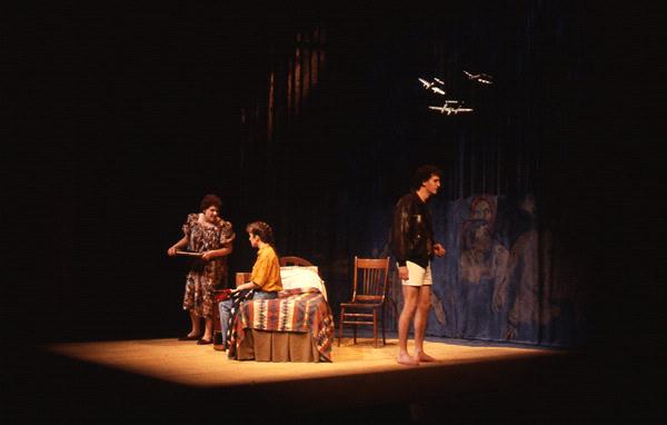 Three actors perform onstage: one stands by a bed, another seated on the bed, and a third at the forefront, wearing a jacket and shorts. The set includes a simple bed and chairs, with airplane models suspended above. Dim lighting creates a moody atmosphere.