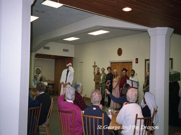 A group of people dressed in medieval costumes perform a scene indoors, watched by an audience seated at tables. The setting resembles a community or dining hall. The audience includes older individuals. Text reads "St. George and the Dragon.