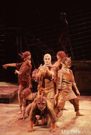 A dramatic stage scene from a production of "Macbeth" showing six performers in earthy brown costumes and masks. They are in dynamic poses on a dimly lit set with a rustic backdrop. The word "Macbeth" is in the bottom right corner.