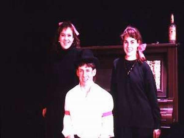 Three people pose in front of a piano. Two women stand on either side, dressed in black with pink bows, and a man kneels in front wearing a white shirt and black hat. A bottle is placed on the piano behind them. The background is dark.