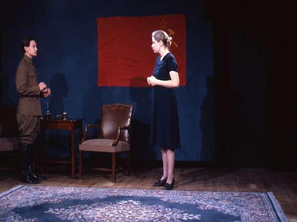 Two people stand in a room with a Soviet flag on the wall. One person, in a military uniform, faces another in a black dress. There are chairs and a small table with glasses nearby, set on a patterned rug.