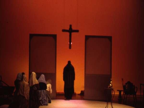 A dimly lit stage with figures in shadowy robes kneeling and standing before a suspended cross. The background is an orange hue with tall rectangular shapes, creating a solemn and atmospheric setting.