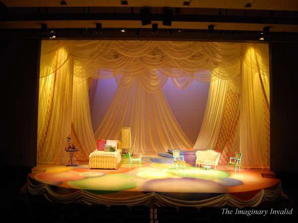 A theater stage set for "The Imaginary Invalid" features draped, elegant curtains and ornate furniture, including a chaise lounge, chairs, and tables. The stage is lit warmly, highlighting colorful circles on the floor.