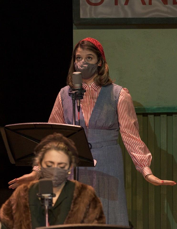 A person in a red hat and mask performs at a vintage-style microphone on stage. They're wearing a striped shirt with a blue dress. Another person in a mask is in the foreground, partially visible, facing downward.