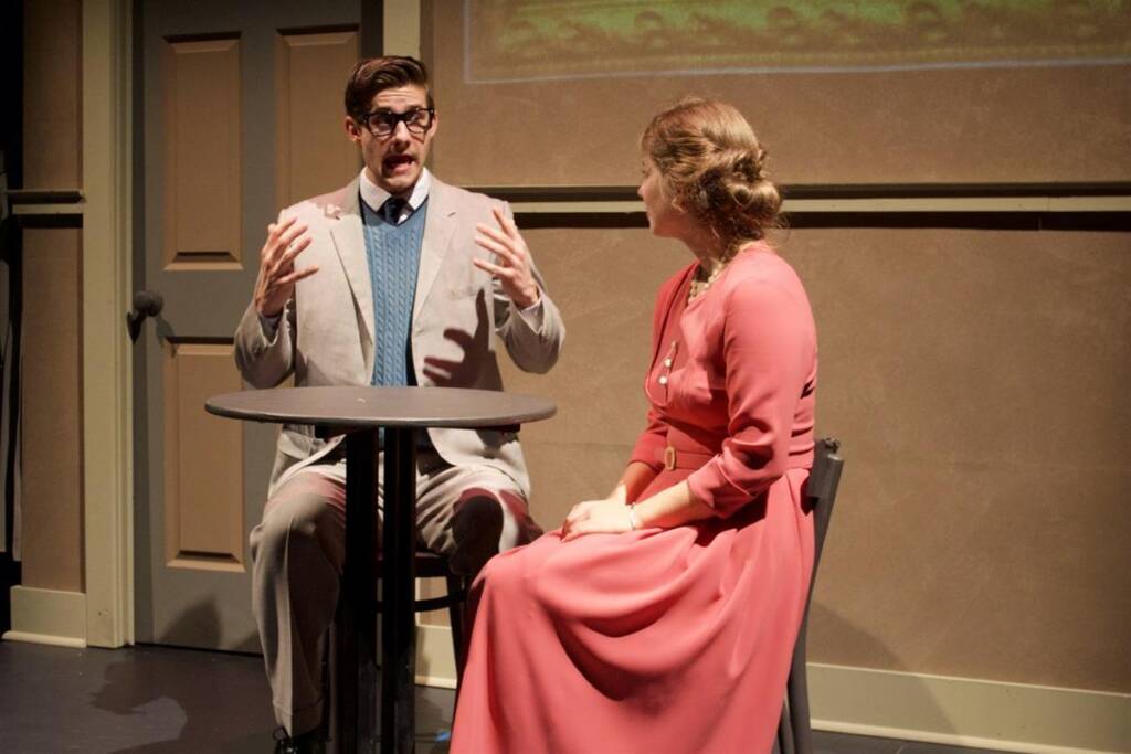 A man and woman sit at a small round table on a stage. The man, wearing glasses and a gray suit, gestures with both hands. The woman in a long pink dress listens attentively. A door and muted background suggest an indoor setting.