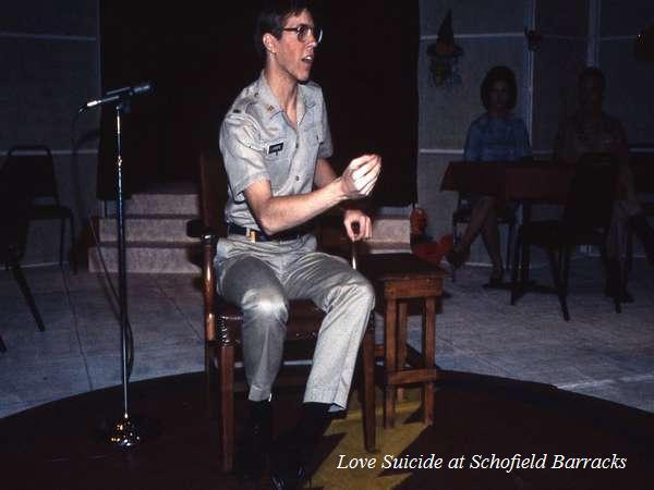 A person in military uniform sits on a wooden chair by a microphone stand. They are gesturing with their hand. The setting appears to be an indoor stage with a few chairs and tables in the background. Text reads "Love Suicide at Schofield Barracks.