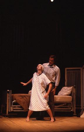 A person in a polka dot hospital gown leans back dramatically while another person stands behind them on a dimly lit stage. A hospital bed is visible in the background, along with a small partition screen.