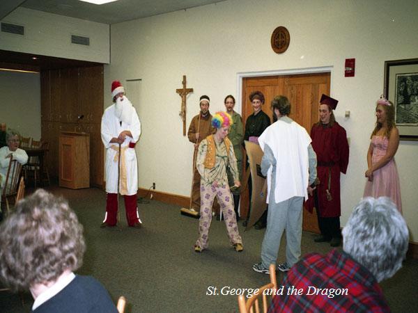 A group of people in costume stand in a room. One person wears a Santa outfit, another a colorful clown wig, others are in medieval attire. They appear to be performing a play. There are seated audience members watching. Text reads "St. George and the Dragon.