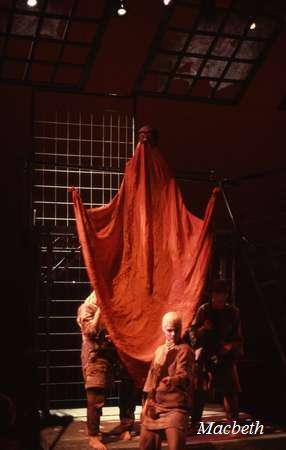 A dramatic stage scene from "Macbeth" featuring actors in medieval costumes. One actor is elevated, draped in a large, flowing red cloth, creating a tall, imposing figure. Other actors stand nearby. Dim lighting enhances the mood. Text reads "Macbeth.