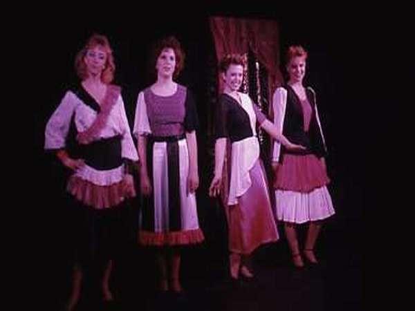 Four women stand on stage in colorful, vintage dresses. They pose confidently with a curtain in the background. The dresses have black, white, and pink accents, and each woman has a distinct hairstyle. The stage setting is dimly lit.