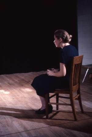 A person with tied-back hair is seated on a wooden chair, facing a dark background. They are wearing a dark outfit and appear deep in thought or focused. The room's wooden floor is illuminated by soft lighting.