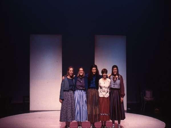 Five people stand on a dimly lit stage, each with arms around one another. They wear long skirts and blouses, and two large rectangular panels are in the background.