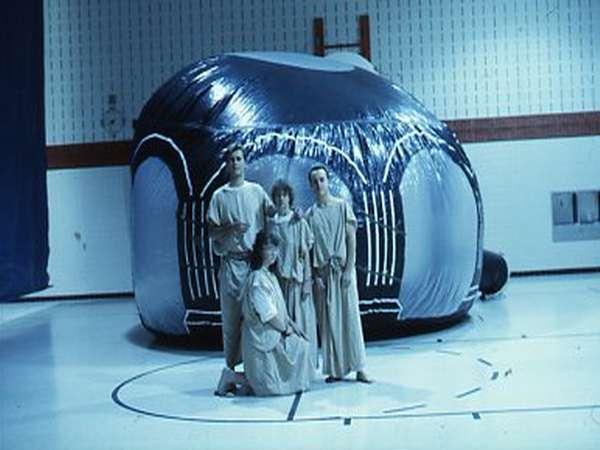 Four people in beige robes stand in front of a large, dome-shaped inflatable structure in a gymnasium. The dome is dark with shiny sections, and the gym has a white floor with sports markings and brick walls.