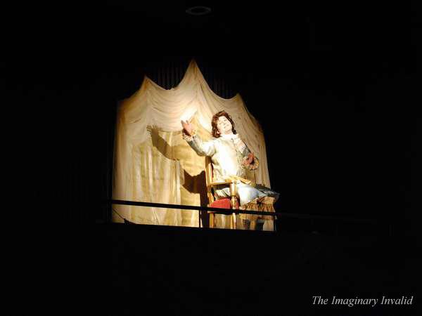 A mannequin dressed in 17th-century style clothing sits on a red chair under a spotlight. It is positioned in front of a draped curtain backdrop. The scene is from "The Imaginary Invalid.