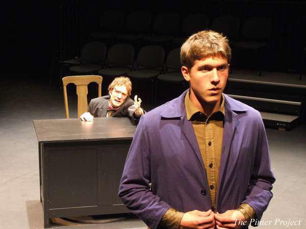 A theater scene with two actors. One man in a blue jacket stands in the foreground, looking determined. Another man is seated behind a desk in the background, raising his hand. The stage is dimly lit with empty seats visible.