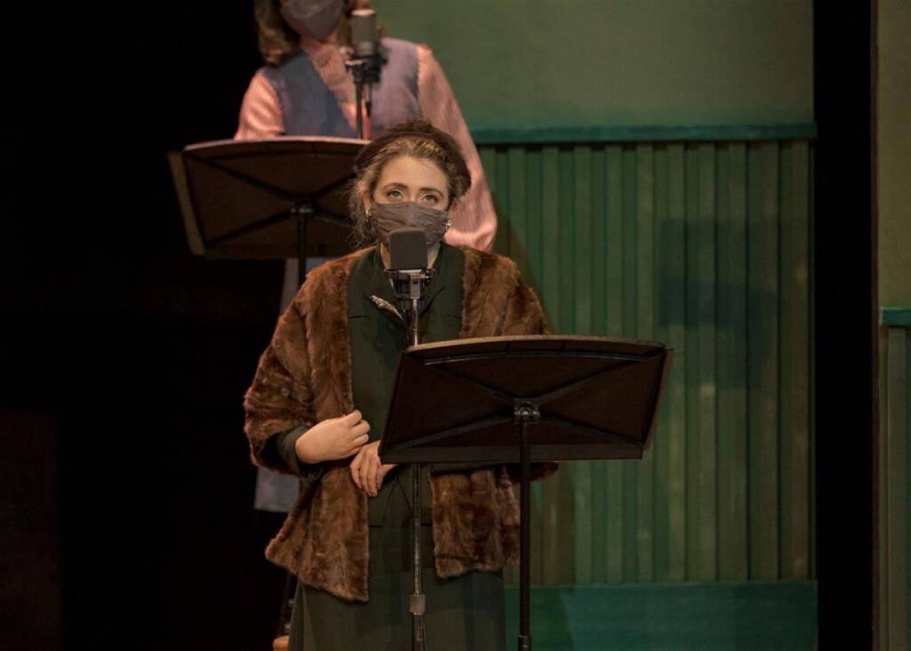 A woman in a fur coat and face mask stands at a microphone with a music stand in front of her. Another person wearing a mask is visible in the background. They appear to be in a dimly lit room.