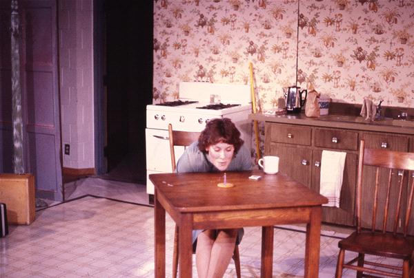 A person leans over a wooden table to blow out a small candle. The kitchen setting includes a stove, cabinets, and a coffee mug on the table. The wallpaper is patterned, and two wooden chairs are placed by the table.