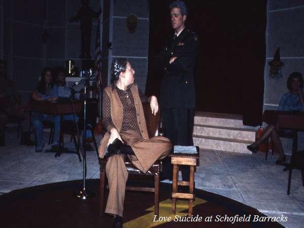 Two actors perform on stage in a scene from "Love Suicide at Schofield Barracks." One actor is seated, wearing a brown suit, and the other stands in military attire with crossed arms. The setting includes tables with people in the background.