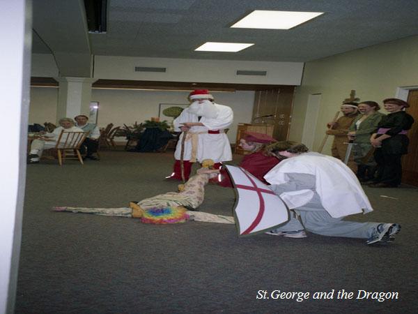 A group of people in costumes performing in a room. One person in a red hat and long robe watches as another in a knight-like outfit kneels over someone in a colorful outfit on the floor. Onlookers are seated and standing nearby. Text reads "St. George and the Dragon.