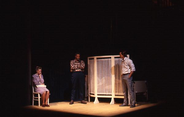 Three people are on a dimly lit stage. A woman sits on a chair to the left, while two men stand talking near a white structure, possibly a room divider. The setting suggests an intimate or serious scene.