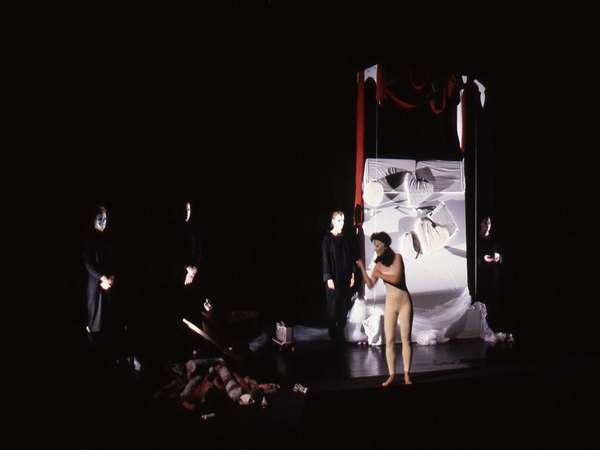 A theatrical performance on a dimly lit stage with abstract props. One performer is in a beige outfit, while others stand in black attire. The backdrop is white with red drapery, creating a dramatic scene with shadows and scattered objects.