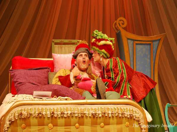 Two performers in vibrant theatrical costumes are on stage. One person, sitting on a lavish bed, has a surprised expression, while the other leans in, appearing to whisper. The backdrop includes rich drapery and a large ornate chair.