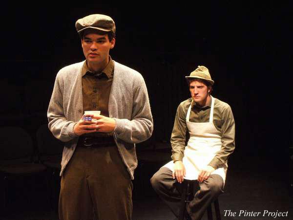 Two actors on stage in a dramatic scene. The foreground actor wears a cap, gray cardigan, and holds an item, looking thoughtful. The background actor sits wearing a cap and apron, appearing contemplative. The setting is dimly lit. Text reads "The Pinter Project.