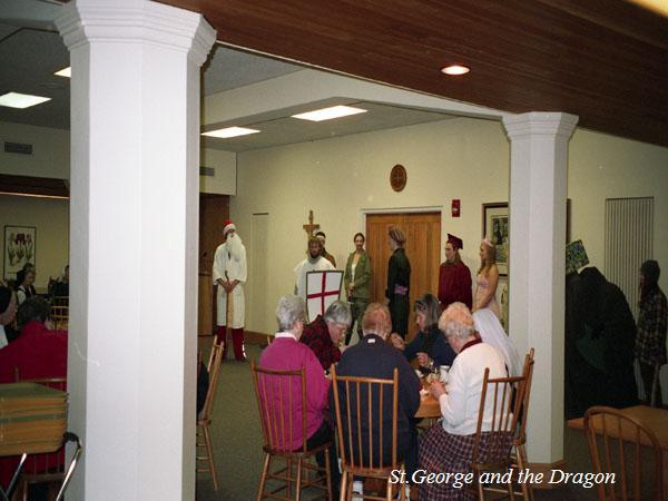 A group of people in costume perform a scene from "St. George and the Dragon" in a room. Some are dressed as knights, and others as medieval characters. An audience sits at tables in the foreground, watching the performance.