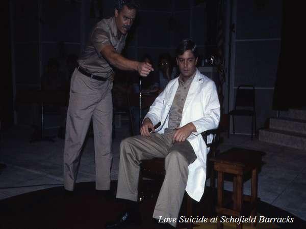 A man in a military uniform points a gun at a seated man wearing a white coat. The scene takes place on a dimly lit stage. The phrase "Love Suicide at Schofield Barracks" is visible at the bottom.