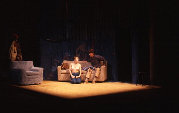 Two actors perform on a dimly lit stage set up as a living room, with one person sitting on a couch and the other kneeling on the floor. A blue armchair, coat, and hat stand are visible in the background.