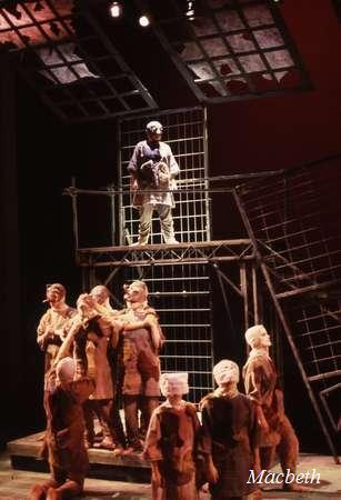 A theatrical scene from "Macbeth" showing an actor standing on a metal platform above the stage, observing a group of performers below. The cast is dressed in dark, rugged costumes, with a rustic backdrop enhancing the dramatic atmosphere.