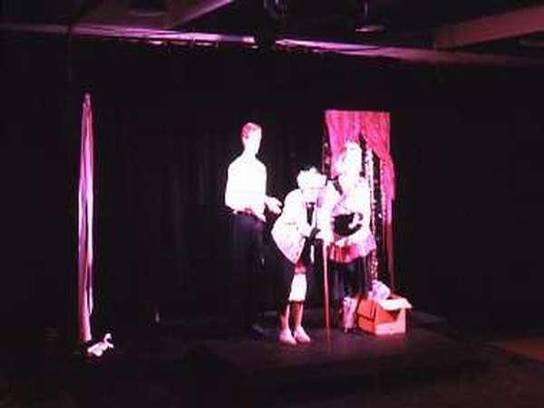 Three actors perform on a dimly lit stage. A person in white uses a cane, another in the middle wears a white shirt, and a third actor in a bright outfit stands beside a prop resembling a box with drapery and streamers hanging.