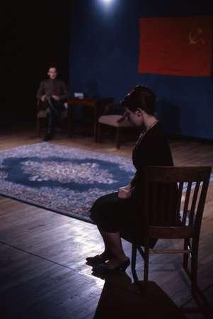 A dimly lit room with two people. One sits on a wooden chair in the foreground facing sideways, while the other sits in the background. A patterned rug lies on the wooden floor, and a red wall hanging is visible.