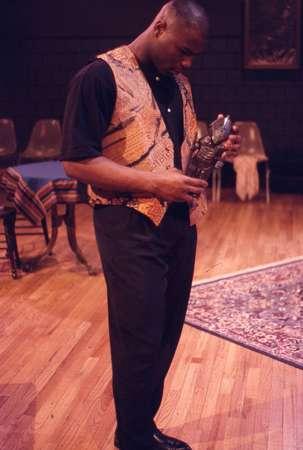 A man stands on a wooden floor, looking down at an object in his hands. He wears a patterned vest over a dark shirt and pants. In the background, there are chairs, a rug, and a table with a colorful cloth.