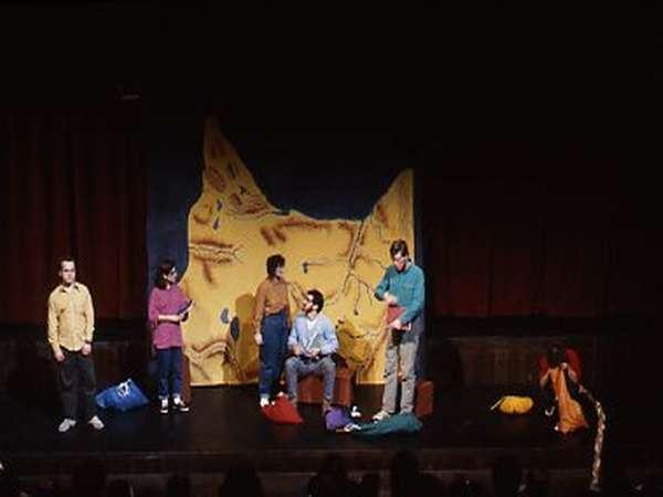 A group of six people on stage performing a scene. They are surrounded by colorful bags and in front of a backdrop painted with abstract shapes. The stage is dimly lit, and the actors have a variety of postures and expressions.