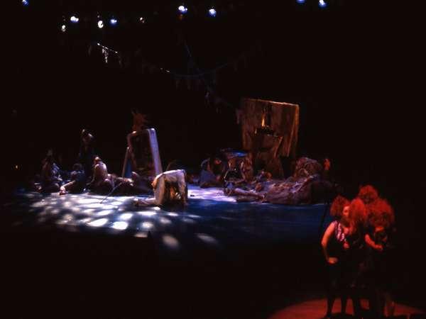 A dimly lit stage with performers in costumes scattered around, some standing and others sitting. The set appears cluttered and abstract, with spotlight patterns on the floor. The overall atmosphere is dark and theatrical.