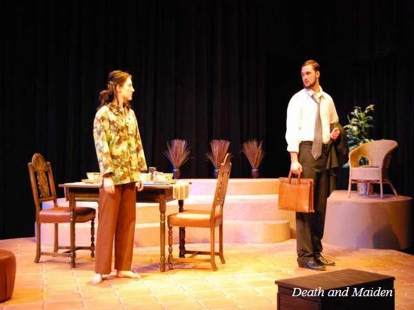 Two actors perform on stage in a scene titled "Death and Maiden." A woman in a patterned blouse faces a man holding a briefcase. They're surrounded by chairs and a table, with decorative plants in the background.