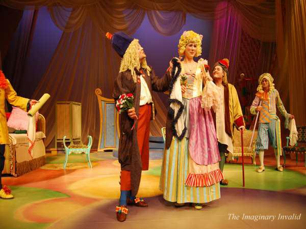 A colorful theatrical scene with actors in period costumes on stage. One actor holds a bouquet, and another, dressed in a striped skirt and yellow wig, interacts with them. The backdrop is draped in elegant fabric. "The Imaginary Invalid" is noted.