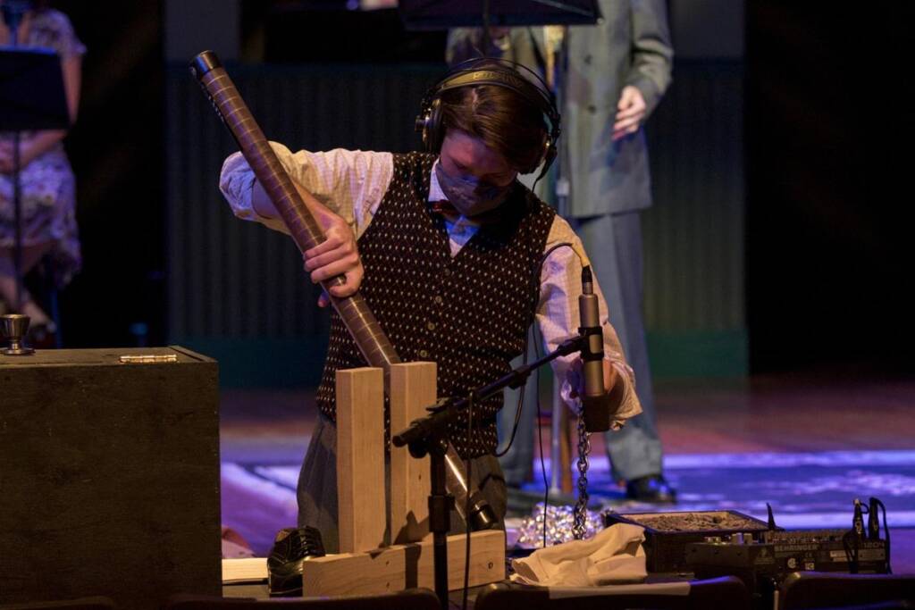 A person in a patterned vest and mask stands on a stage, holding a large wooden tube near a microphone. They are surrounded by sound equipment and wearing headphones, appearing to create sound effects.