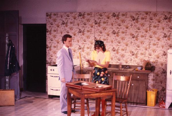 A man and woman stand in a vintage kitchen with floral wallpaper. The man, in a light suit, looks at the woman, who is wearing a yellow top and floral skirt, holding a notepad by a wooden table with chairs. A stove and fridge are in the background.