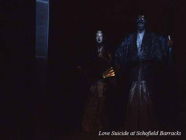 A dark stage features two individuals in traditional Asian costumes and masks, illuminated subtly. One holds a fan, while the other, taller figure wields a staff. Text in the corner reads, "Love Suicide at Schofield Barracks.