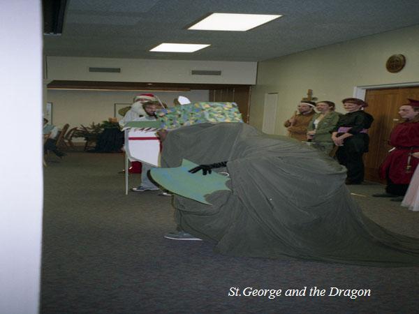 A group of people are dressed in costumes indoors. One person is in a white robe, others stand at the side wearing various outfits, including one holding a sword. The scene likely depicts a performance of "St. George and the Dragon.