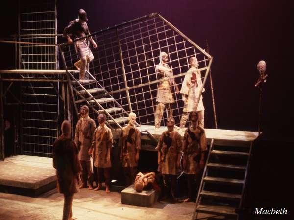 A theatrical scene from "Macbeth" featuring actors in a dimly lit stage set with metal grids and steps. Performers wear tattered costumes, carrying poles, and one actor is elevated on the stairway. The atmosphere is dark and intense.